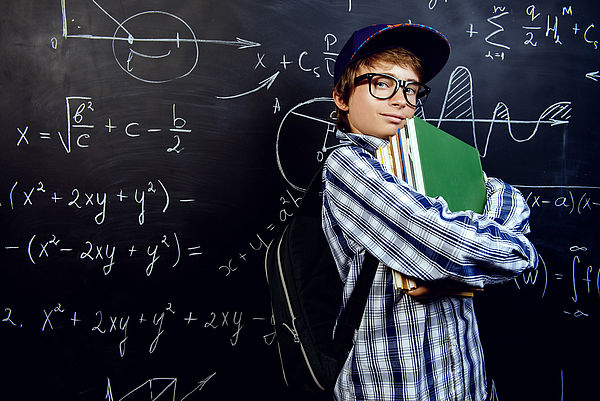 Ein Schüler steht vor einer Tafel, die mit Formeln voll geschrieben ist. Er hält Bücher in seiner Hand und lächelt selbstbewusst