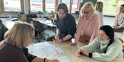 4 Personen stehen bzw. sitzen und schauen auf auf dem Tisch liegende Unterlagen